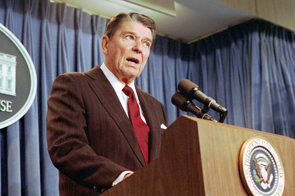 President Reagan, in a brown pinstripe suit and red tie, speaks at a lectern bearing the presidential seal, with a blue curtain and part of The White House sign behind him.