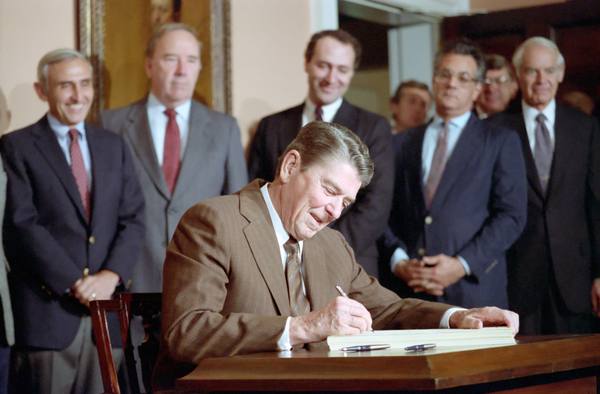 President Reagan leaning over a wooden desk to sign a thick bill with a pen while several men in suits stand smiling behind him.
