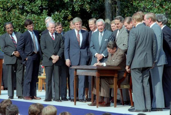 President Reagan seated at a small wooden desk on an outdoor stage, signing a bill while a group of men in suits stands close around him.
