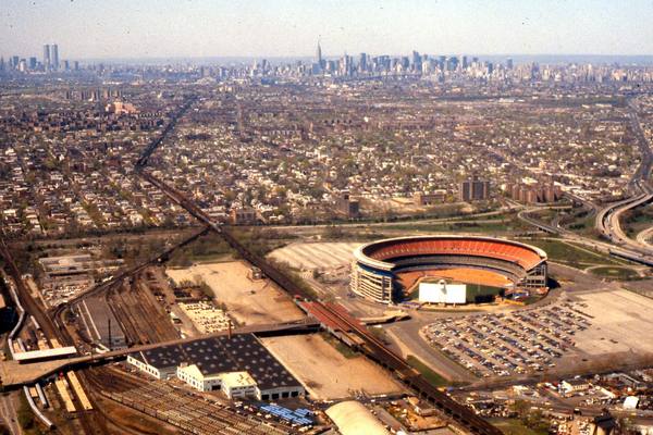 From the air, the round orange-seated Shea Stadium sits beside a large parking lot and rail yards in Queens, with the Manhattan skyline and the twin World Trade Center towers on the horizon.