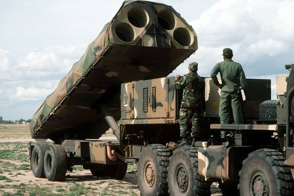 A camouflage-painted truck and trailer carrying a launcher tilted upward so that its four large round launch tubes face the viewer, with two uniformed servicemen standing on the truck, seen from behind.