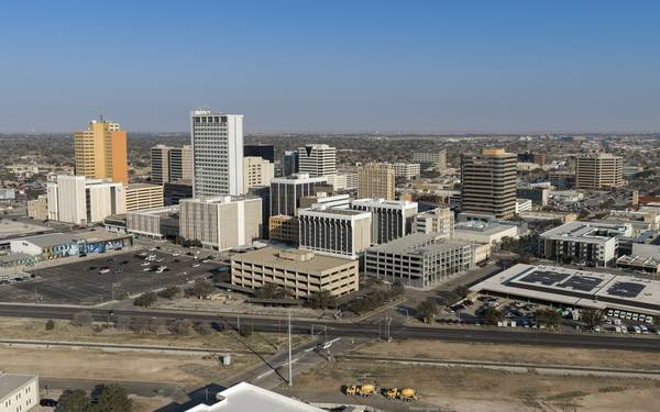 An elevated view of downtown Midland, Texas, with office towers and parking garages under a clear blue sky, a road and railway tracks in the foreground and flat plains stretching to the horizon.