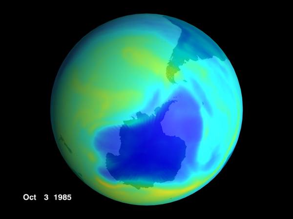 A globe seen from above the South Pole, coloured blue, green, yellow and cyan, with a deep blue patch of very low ozone covering Antarctica and a small white date label reading Oct 3 1985 at lower left.