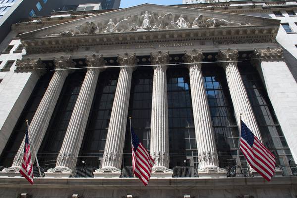 Looking up at the marble columns, sculpted pediment and lettering of the New York Stock Exchange building, with three American flags hanging in front.