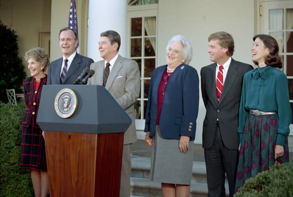 Six smiling people stand beside a wooden podium bearing the presidential seal outside a white-columned building: a woman in a red and black plaid suit, a man in a gray suit, a man in a tan jacket at the microphones, a white-haired woman in a navy jacket, a man in a dark suit with a red striped tie, and a woman in a green blouse.