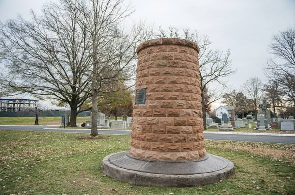 A round tower of rough-cut red sandstone blocks with a small dark plaque, standing on a low stone base inscribed with text, among bare trees and headstones in a cemetery.