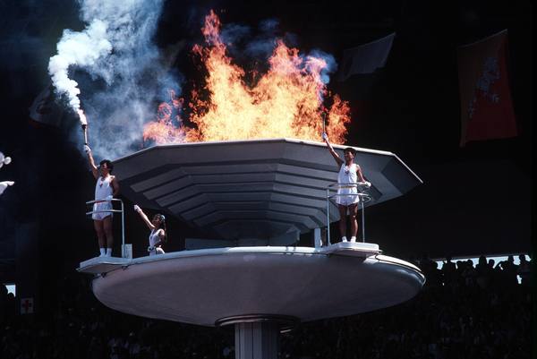 Two men and a woman in white sleeveless outfits stand on the white platform of a large octagonal cauldron, two of them raising torches, one trailing white smoke, as tall flames rise from the bowl against a dark stadium background.