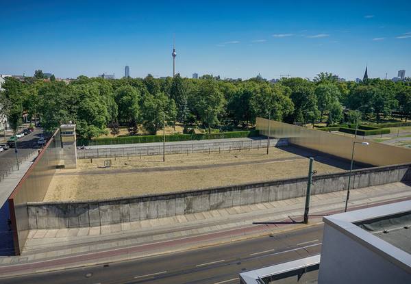 Two long stretches of gray concrete wall with a small watchtower and a strip of bare sandy ground between them, framed by tall brown and gold-toned steel panels, with green trees and the Berlin TV tower beyond under a blue sky.