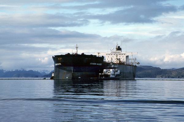 A large dark-hulled oil tanker with the name Exxon Valdez on its bow and a white superstructure sits on calm water under a cloudy sky, with a small tug alongside and snow-capped mountains behind.