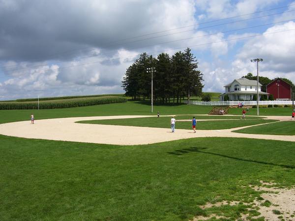 A baseball diamond with a few small figures playing on it, a white farmhouse and red barn in the distance, a stand of tall evergreen trees and a field of corn, under a dramatic cloudy sky.