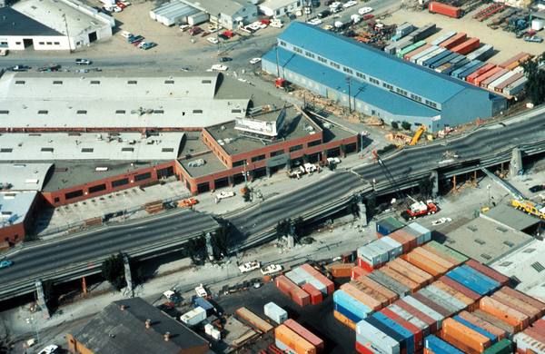 An aerial view of an elevated freeway with a buckled, collapsed stretch running past warehouses and rows of colourful shipping containers, with a crane and a few small vehicles and workers nearby.