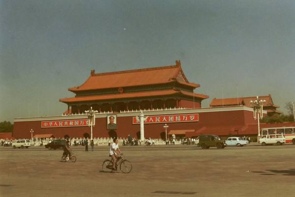 A red-walled gate tower with two tiers of orange tile roofs and a portrait above the central archway, with red banners on the wall, a white marble column, and a few cyclists, cars and a bus on the wide paved square in front.