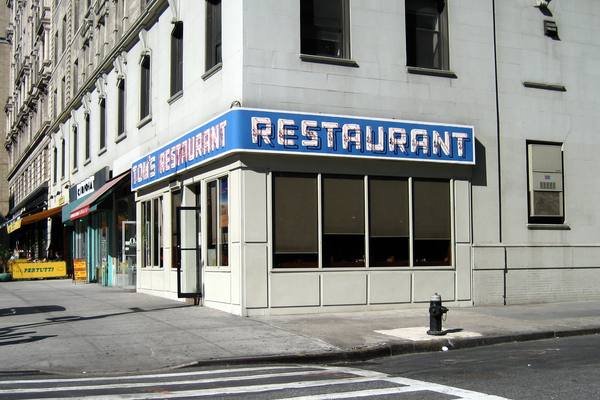 The corner of a diner with a blue sign reading Tom's Restaurant in neon lettering above large windows, on a sunny city street corner with a fire hydrant.