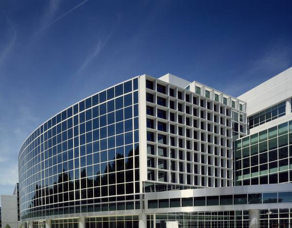 The curved glass-and-white-panel facade of a large modern office-style building under a deep blue sky with thin streaks of cloud.
