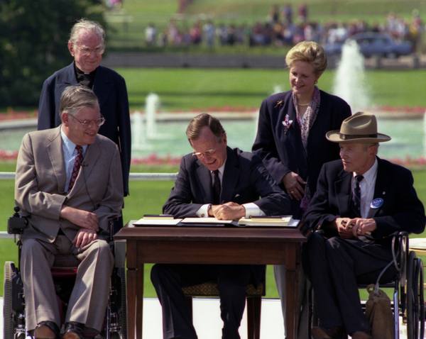 President Bush seated at a small table signing a document on a lawn, with a man in a wheelchair on the left, another man in a wheelchair wearing a wide-brimmed hat on the right, and a priest and a woman standing behind.