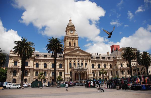 A cream-coloured city hall with a central clock tower, palm trees and market stalls in the square in front, under a blue sky with a bird in flight.