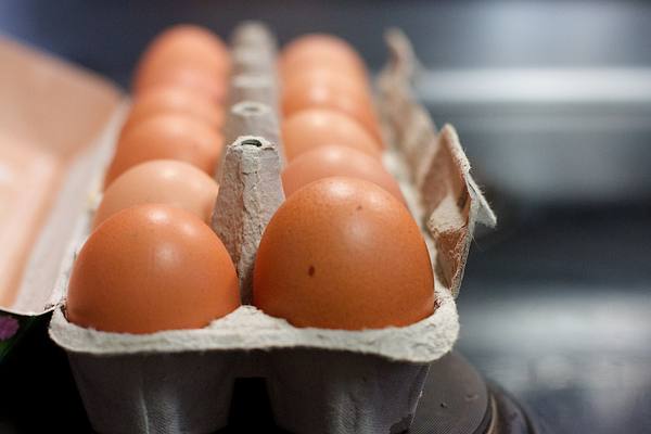 Brown chicken eggs in a grey cardboard carton, two sharp eggs in the foreground and more eggs softly blurred behind them against a dark background.