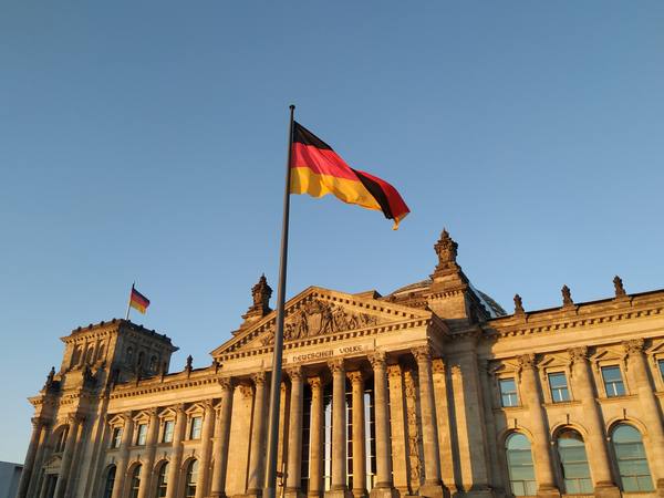 A black-red-gold German flag on a tall pole in front of the stone Reichstag building, in warm evening light against a clear blue sky.