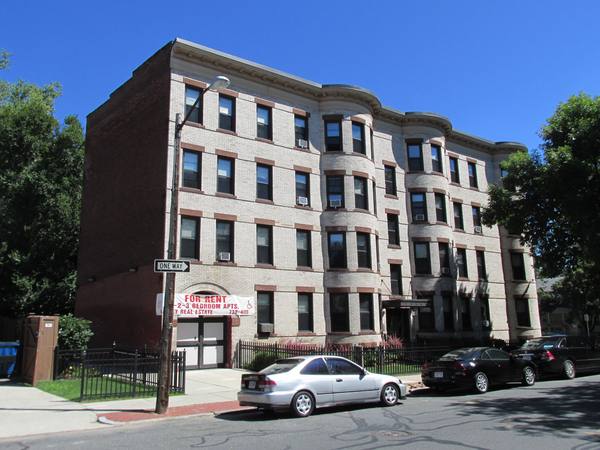 A four-storey cream-brick apartment building with curved bay windows, a red-and-white For Rent banner over the doorway and cars parked along the street.