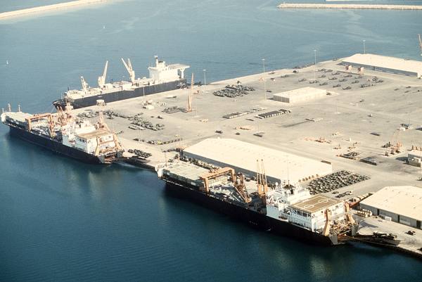 Aerial view of a large concrete quay with three dark-hulled cargo ships moored along it, military vehicles and cargo stacked on the quay, rows of parked helicopters near the far warehouse, and large warehouses on the right.