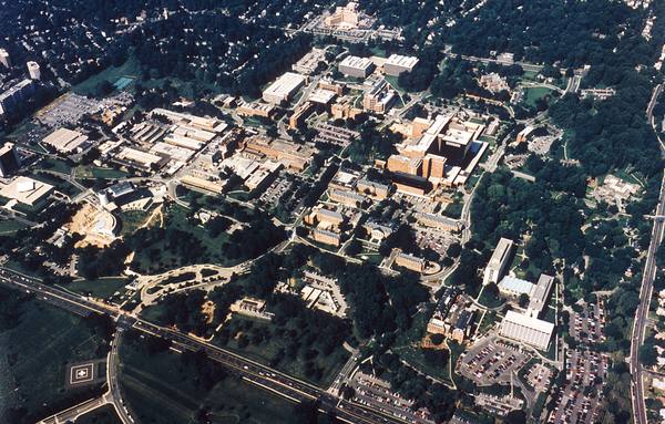 An aerial view of a wooded suburban campus of low and mid-rise buildings, roads and parking lots, with a helicopter landing pad at the lower left.