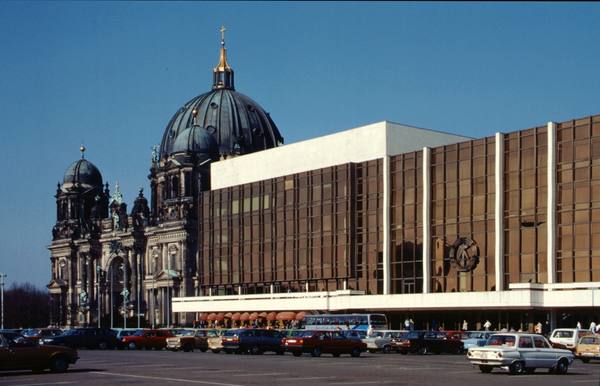 A long modern building with bronze-tinted glass walls and a white upper block beside the dome of Berlin Cathedral, with cars parked in the square in front.