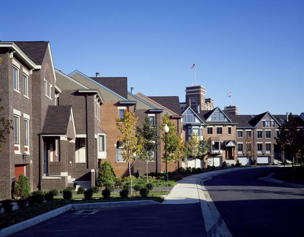 A curving residential street with a sidewalk, young trees and rows of brick houses with garages under a clear blue sky, and a brick tower with flags in the distance.