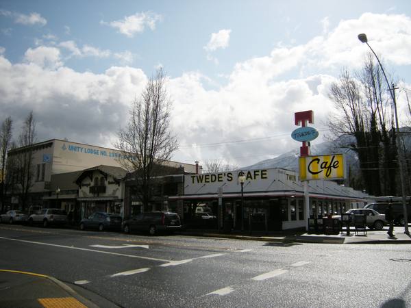 A street corner in a small mountain town with a low white roadside cafe lettered Twede's Cafe, a tall sign reading Cafe, bare trees, parked cars, a wet road and a cloudy sky above a mountain slope.