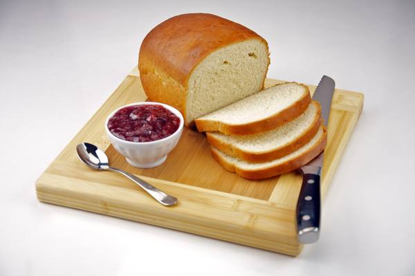 A golden-crusted loaf of white bread with three slices cut off, beside a small white bowl of jam, a spoon and a bread knife on a bamboo cutting board.