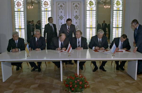 Officials in dark suits seated at a long white table signing documents in a room with tall stained-glass windows, small national flags on the table and a bouquet of red flowers in front.