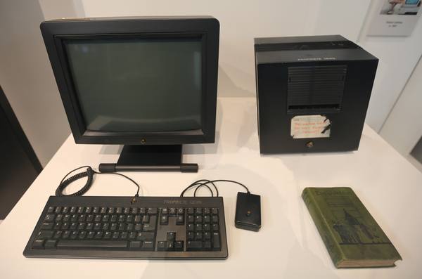 A black NeXT computer cube with a handwritten warning label sits beside a NeXT monitor, keyboard, mouse and an old green book on a white display table.