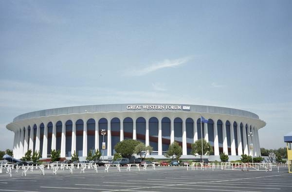 A large round arena with white arched pillars and a 'Great Western Forum' sign on its roof edge, above a mostly empty parking lot under a pale blue sky.