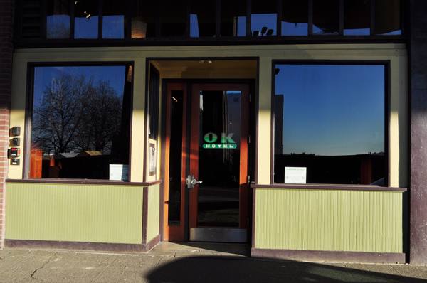The street-level front of a small hotel building with a wooden door marked OK Hotel, two large windows reflecting bare trees and sky, and green wood-panelled lower walls.