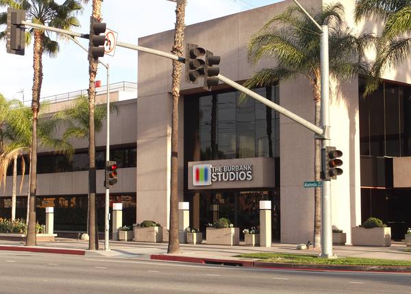 The street front of a large pale stone studio building with a Burbank Studios sign, palm trees, traffic signals and an empty road in front.