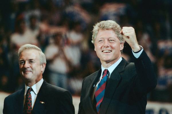 Bill Clinton smiling and raising a clenched fist at a rally, beside a smiling older man in a suit, with a blurred crowd behind them.