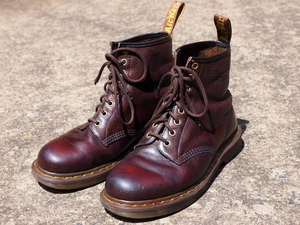 A pair of scuffed oxblood-brown leather lace-up boots with yellow stitching, yellow pull tabs and thick soles, standing on a pale concrete surface.