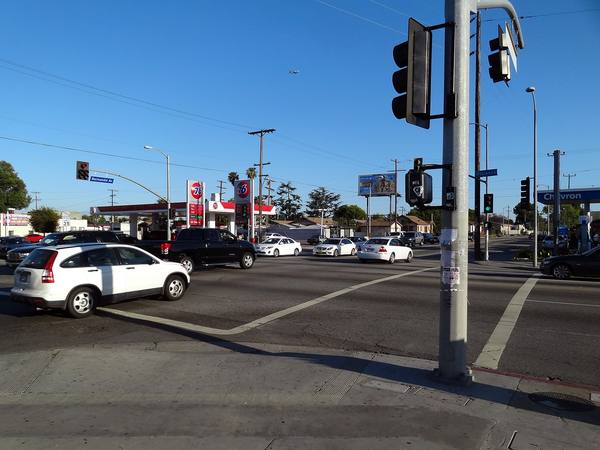 A South Los Angeles street corner under a clear blue sky, with a white SUV and other cars waiting at the intersection, a traffic-signal pole on the right, street signs reading Normandie and Florence, and two gas stations in the background.