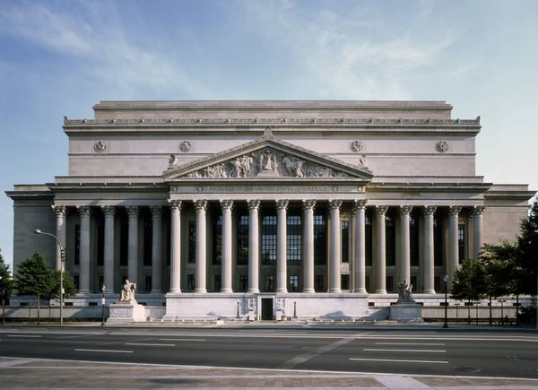 The columned neoclassical front of the National Archives Building under a pale blue sky, with the words Archives of the United States of America across the entablature.