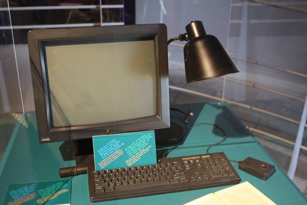 A black NeXT computer with a flat-faced monitor, keyboard and mouse on a teal display table, with a black desk lamp, a small teal information card standing in front of the screen and an open book at the lower right.