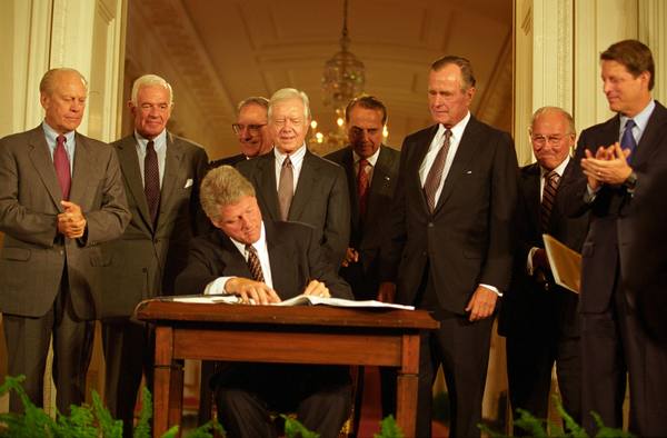 President Bill Clinton, seated at a small wooden desk, signs a large bound document while eight men in suits, including three former presidents and the vice president, stand behind him in a white-columned room with a chandelier.