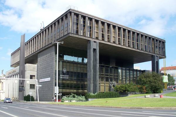 A dark, boxy modernist building raised on large pillars, with rows of tall windows and the words Národní Muzeum on its glass front, beside a road and a grass lawn under a blue sky with clouds.