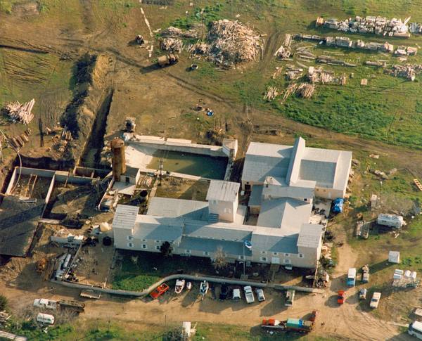 An aerial view of a cluster of pale-roofed buildings, a rusty water tower and a pool courtyard on open farmland, with vehicles parked along a dirt road below and piles of lumber in the fields.