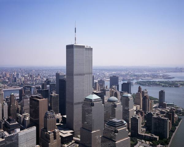 The two tall twin towers of the World Trade Center rising above lower Manhattan, with a river and a low city stretching into the hazy distance under a pale blue sky.
