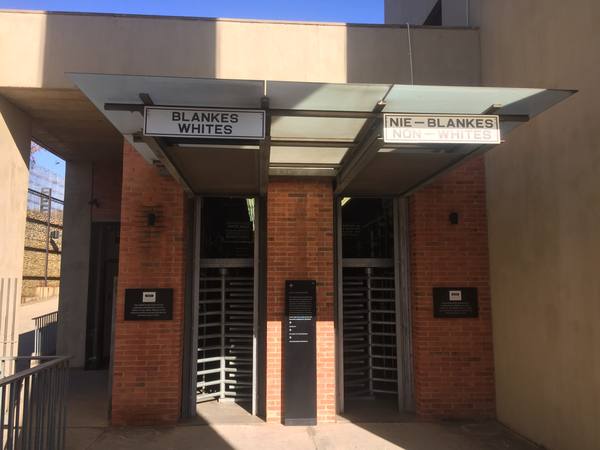 A red-brick museum entrance with two turnstile gates under a glass canopy; the left sign reads 'Blankes / Whites' and the right sign reads 'Nie-Blankes / Non-Whites', with a dark plaque beside each gate explaining that the visitor's ticket decides which to use.