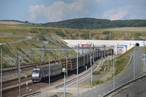 A silver electric locomotive numbered 9705 pulls a silver coach and a long train of tall lattice-sided wagons past a white tunnel portal painted with the words Euro Tunnel, set into a green chalk hillside, with an empty road curving up the right-hand side.