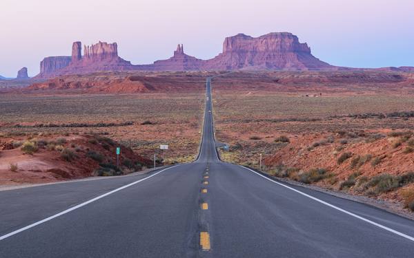A straight two-lane highway runs toward distant red sandstone buttes under a pale pink and violet dawn sky, with a small green mile marker beside the road at left.