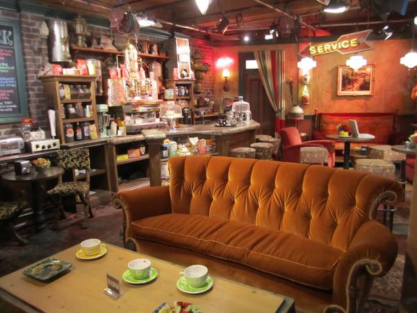 A coffeehouse set with a tufted orange velvet sofa and a wooden coffee table with three cups in the foreground, and a counter with espresso machines, brick walls and a neon Service sign behind.