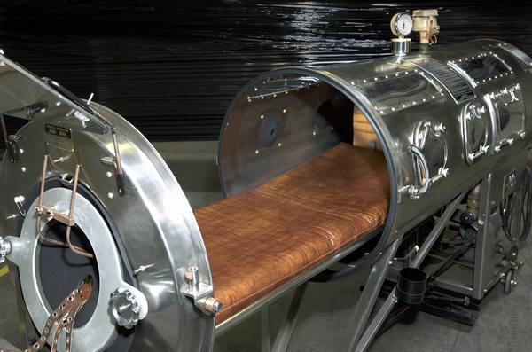 A polished metal iron lung with its end opened, showing a brown padded sliding bed and a pressure gauge on top, in front of a black plastic-wrapped backdrop.