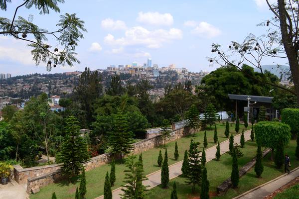 Landscaped memorial gardens with a paved path, a stone retaining wall and conifer trees in the foreground, a low open-sided building at the right, and the hills and skyline of Kigali beyond under a blue sky with white clouds.