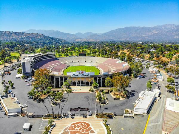 An aerial view of the Rose Bowl stadium with its green pitch, maroon seating and 'Rose Bowl' sign over the tunnel entrance, a large rose mosaic on the plaza in the foreground, car parks and trees around it and a mountain range under a clear blue sky.